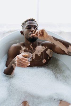 Foto de Hombre afroamericano con vitiligo fumando cigarro y sosteniendo whisky mientras toma un baño en casa - Imagen libre de derechos