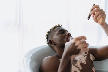 Young african american man with vitiligo looking at cigar and holding glass while taking bath at home 