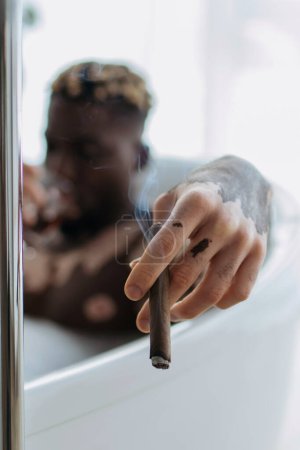 Téléchargez les photos : Homme afro-américain flou avec du vitiligo tenant un cigare tout en prenant un bain à la maison - en image libre de droit