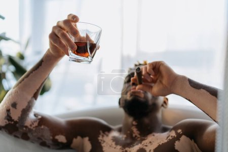 Foto de Hombre afroamericano borroso con vitiligo fumando cigarro y sosteniendo whisky mientras toma un baño en casa - Imagen libre de derechos