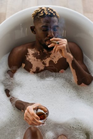 Vue du dessus de l'homme afro-américain avec du cigare fumant du vitiligo et tenant du whisky dans un bain avec mousse 