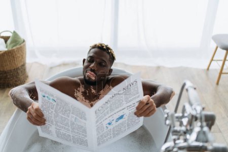 Joven afroamericano con vitiligo leyendo periódico económico mientras toma baño en casa 