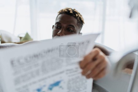 Foto de Hombre afroamericano con vitiligo leyendo un periódico borroso mientras toma un baño en casa - Imagen libre de derechos
