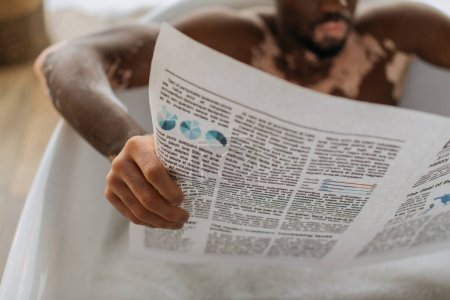 Vista recortada de un hombre afroamericano borroso con vitiligo leyendo el periódico mientras toma un baño por la mañana 