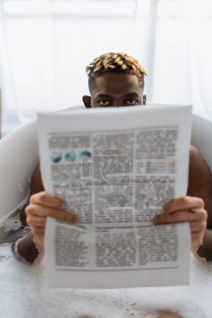 African american man with vitiligo covering face with newspaper in bath with foam at home 