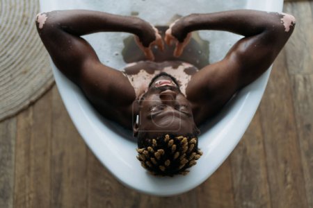 Téléchargez les photos : Vue aérienne de l'homme afro-américain avec vitiligo écouter de la musique et prendre un bain à la maison - en image libre de droit