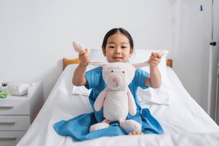 Foto de Asiático chica en hospital bata sonriendo en cámara mientras jugando con juguete conejito en cama en clínica - Imagen libre de derechos