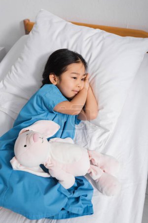 Foto de Alto ángulo vista de soñador asiático niño acostado en hospital cama cerca juguete conejito y mirando lejos - Imagen libre de derechos