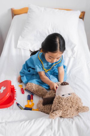 high angle view of asian girl examining teddy bear with toy stethoscope while playing on bed in hospital