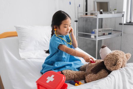 Foto de Poco asiático chica examinando teddy oso con juguete estetoscopio en hospital cama - Imagen libre de derechos