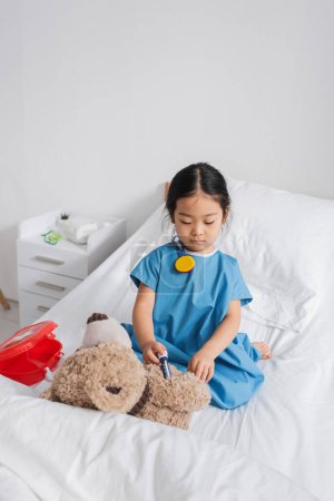 asiatisches Kind im Krankenhauskleid injiziert Teddybär mit Spielzeugspritze beim Spielen auf dem Bett in Klinik