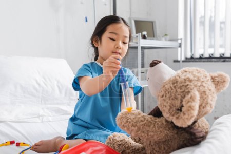 little asian child in hospital gown doing injection to teddy bear with toy syringe 