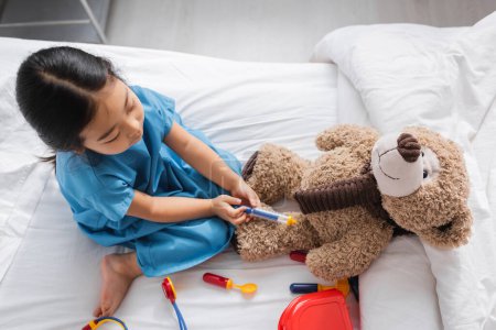 top view of asian child playing on hospital bed and doing injection to teddy bear with toy syringe