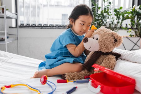 Asiatisches Kind im Krankenhauskittel untersucht Teddybär mit Spielzeug-Otoskop auf Bett in Kinderklinik