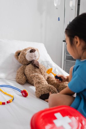 Asiatisches Kind untersucht Teddybär mit Reflexhammer auf Krankenhausbett