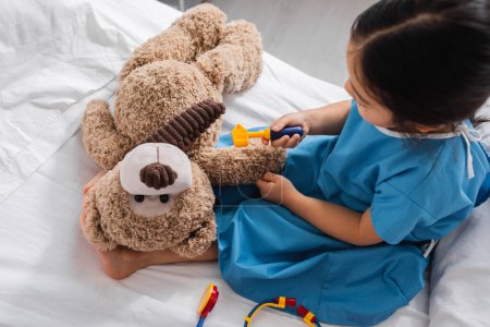 top view of asian kid in hospital gown holding toy reflex hammer near teddy bear while playing in clinic