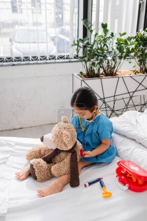 asian child in hospital gown examining teddy bear with toy stethoscope on bed in clinic