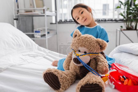 asian girl looking at camera while playing with teddy bear and toy medical equipment on hospital bed