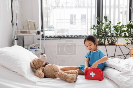 Asiatin öffnet Spielzeug-Verbandskasten in der Nähe von Teddybär auf Bett in moderner Kinderklinik