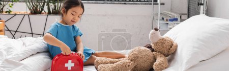 Photo for Asian child opening toy first aid kid near teddy bear on hospital bed, banner - Royalty Free Image