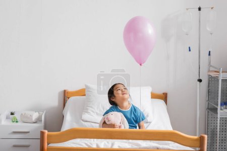 joyful asian girl sitting on hospital bed with toy bunny and looking at festive balloon