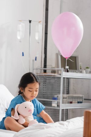 kleines asiatisches Kind spielt mit Spielzeughasen, während es auf dem Krankenhausbett in der Nähe eines festlichen Ballons sitzt