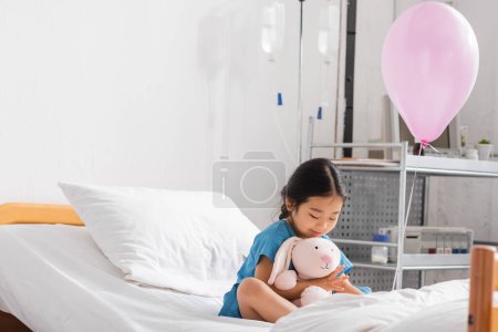 cheerful asian girl playing with toy bunny near festive balloon on hospital bed