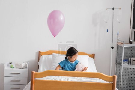 little asian girl playing with toy bunny on bed under festive balloon in hospital ward