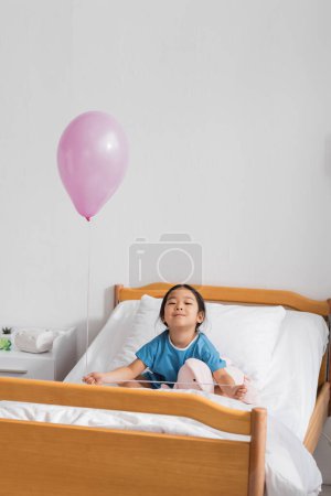Foto de Feliz asiático chica con festivo globo y juguete conejito sentado en hospital cama y sonriendo a cámara - Imagen libre de derechos