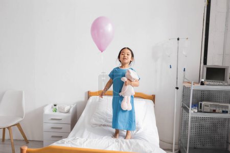 full length of cheerful asian child standing on hospital bed with toy bunny and festive balloon