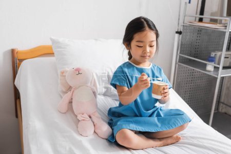 pleased asian girl holding tasty yogurt while sitting on hospital bed near toy bunny