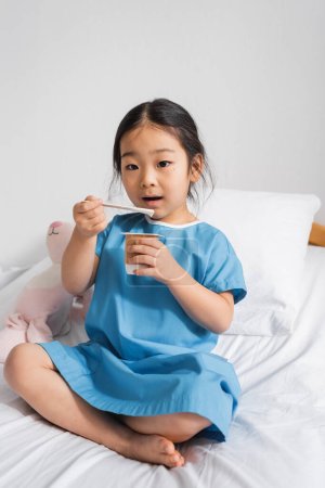 asiatisches Mädchen isst köstlichen Joghurt und schaut beim Frühstück im Krankenhaus in die Kamera