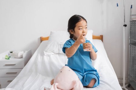 asiatisches Kind isst köstlichen Joghurt und schaut in der Nähe von Spielzeughase auf Krankenhausbett weg