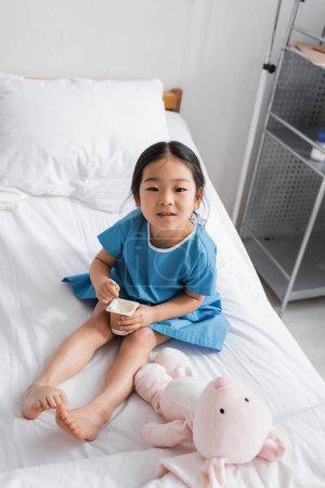 positive asian girl holding delicious yogurt and smiling at camera in hospital ward