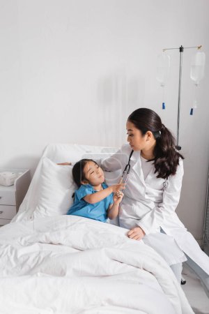 Neugieriges asiatisches Mädchen berührt Stethoskop in der Nähe von Arzt auf Krankenhausstation