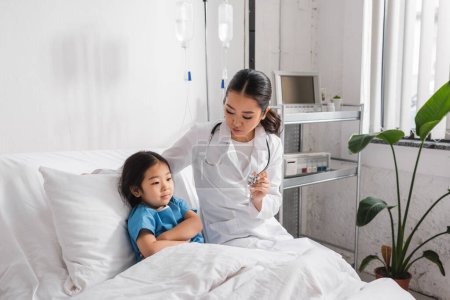 Foto de Joven médico mostrando estetoscopio a asiático chica sentado en hospital cama con los brazos cruzados - Imagen libre de derechos