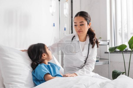 young asian pediatrician with stethoscope looking at girl on bed in hospital ward