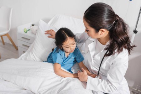 curious asian child touching stethoscope near doctor on bed in hospital 