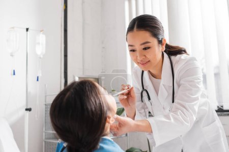 sonriente asiático médico celebración lengua depresor mientras examinar poco paciente en hospital sala