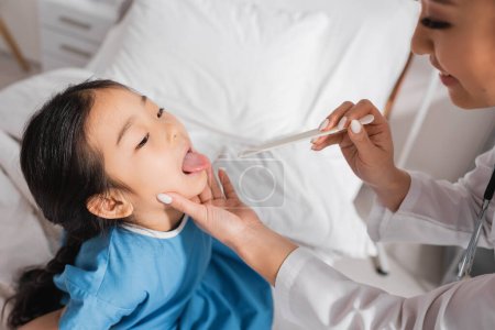 asiático chica apertura boca cerca sonriente médico con lengua depresor en hospital sala