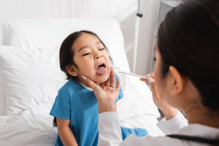 Foto de Borrosa pediatra celebración lengua depresor cerca asiático chica apertura boca en clínica - Imagen libre de derechos