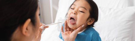 asiatisches Kind mit offenem Mund in der Nähe von Arzt mit Zungendepressor auf verschwommenem Vordergrund auf Krankenhausstation, Banner