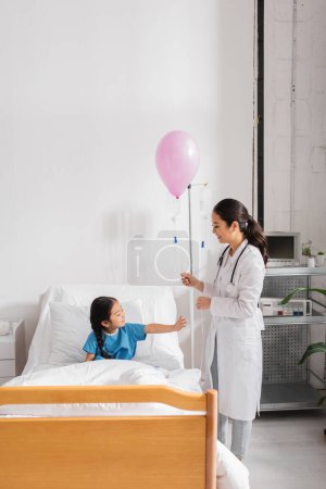 Foto de Sonriente médico en blanco abrigo de pie con festivo globo cerca de asiático chica en hospital cama - Imagen libre de derechos