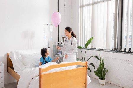lächelnder asiatischer Arzt mit festlichem Luftballon in der Nähe eines Mädchens, das auf einer modernen Krankenhausstation auf dem Bett sitzt