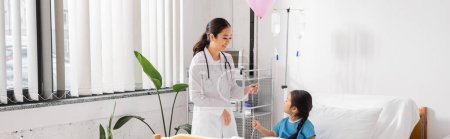 Téléchargez les photos : Heureux asiatique médecin en blanc manteau tenant ballon festif près de l'enfant dans moderne clinique pédiatrique, bannière - en image libre de droit