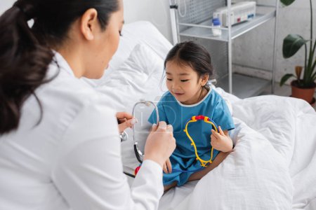 blurred doctor showing stethoscope to positive asian girl sitting with toy on hospital bed