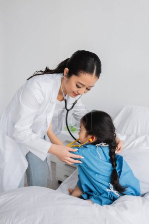 smiling asian pediatrician with stethoscope examining child in hospital gown sitting on bed in clinic