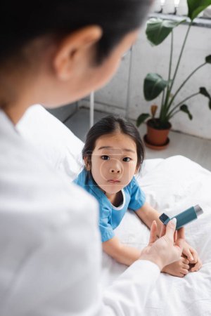 Foto de Pediatra borrosa mostrando inhalador a niño asiático sorprendido en la sala de hospital - Imagen libre de derechos