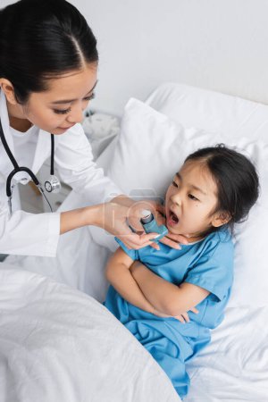 Junge Kinderärztin hält Inhalator nahe asiatischem Mädchen mit offenem Mund und verschränkten Armen auf Krankenhausbett