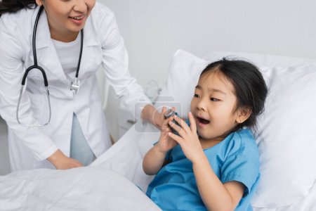 little asian girl with open mouth holding inhaler near doctor in pediatric clinic
